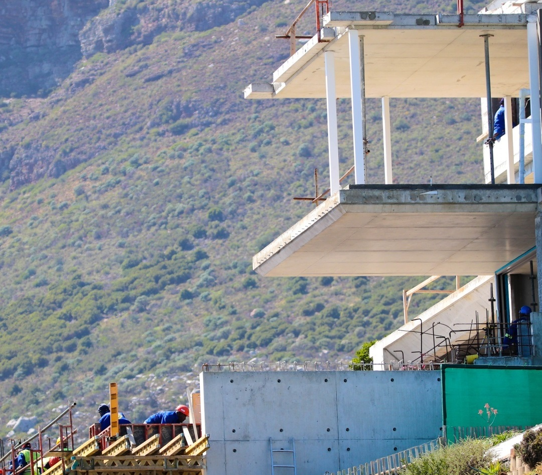 A multi-story residential building under construction against a mountain backdrop, showcasing a long, unsupported post tension slab cantilever balcony with temporary shoring props.